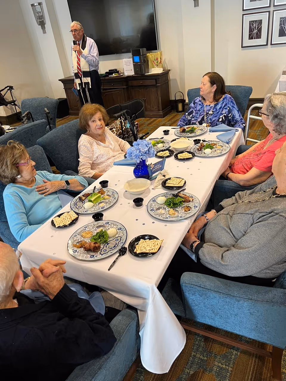 A group of elderly residents seated around a table in a dining area with plates of food while a man speaks into a microphone near a TV.