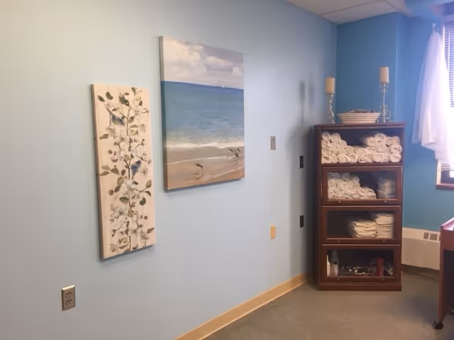 Interior room with light blue walls, two framed paintings and a wooden shelf filled with rolled towels near a window.
