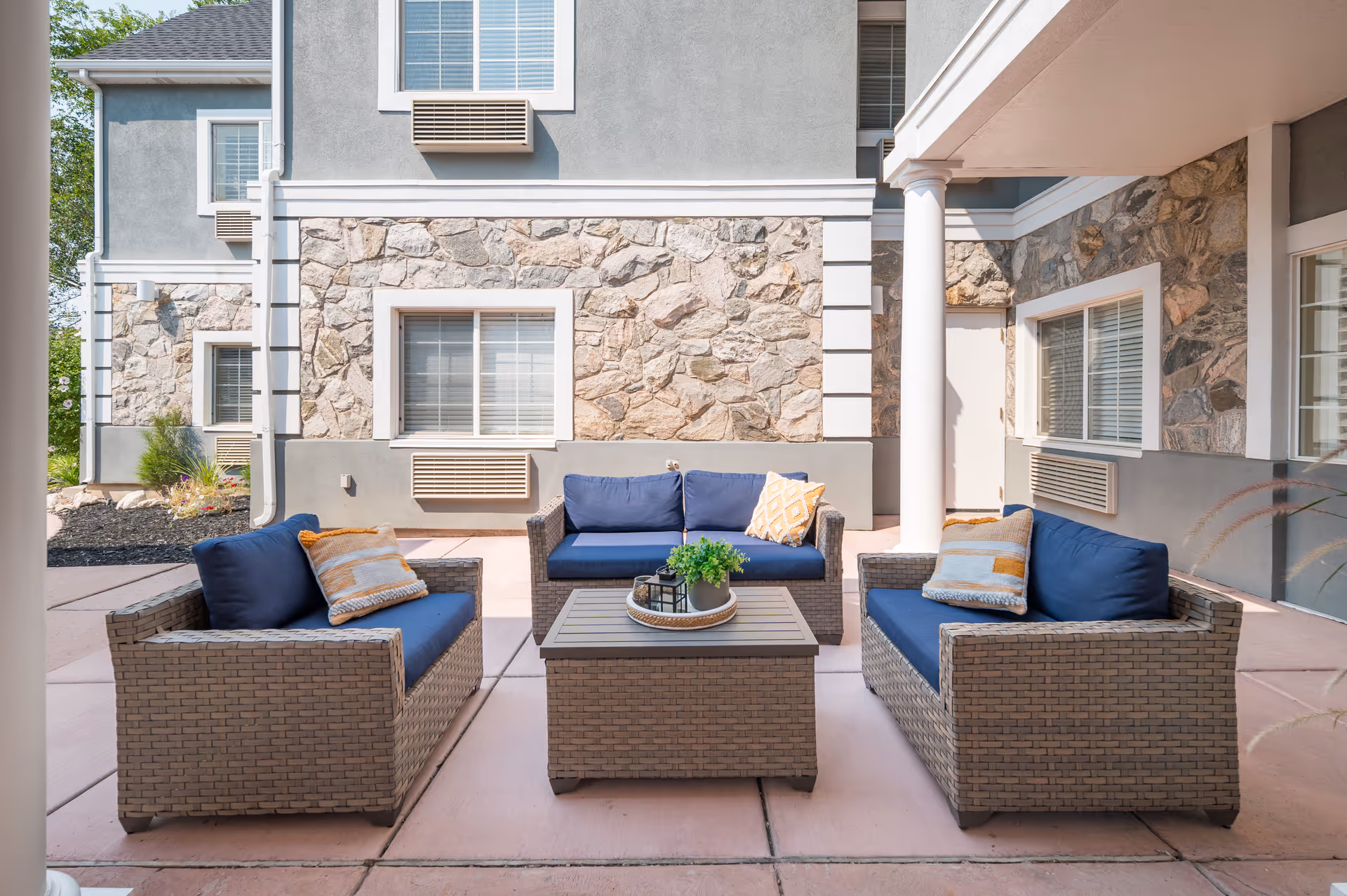 Outdoor patio area with three wicker sofas featuring blue cushions and decorative pillows arranged around a square wicker coffee table with a plant and lantern on top. The background shows a stone and stucco building exterior with windows and air conditioning units.