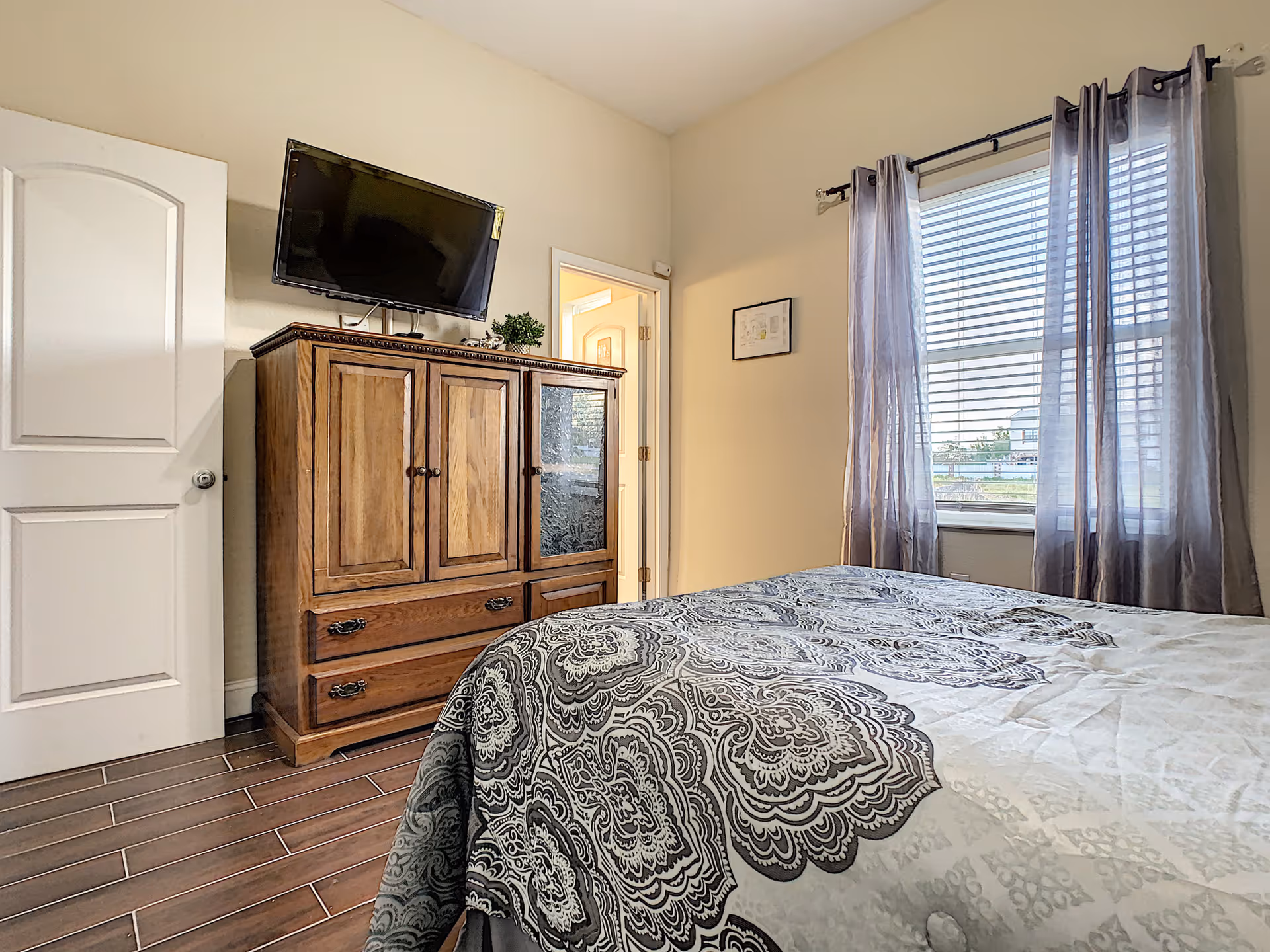 A bedroom with a bed covered in a patterned gray and white comforter, a wooden dresser with a flat-screen TV mounted above it, a window with sheer gray curtains, and a door leading to another room.