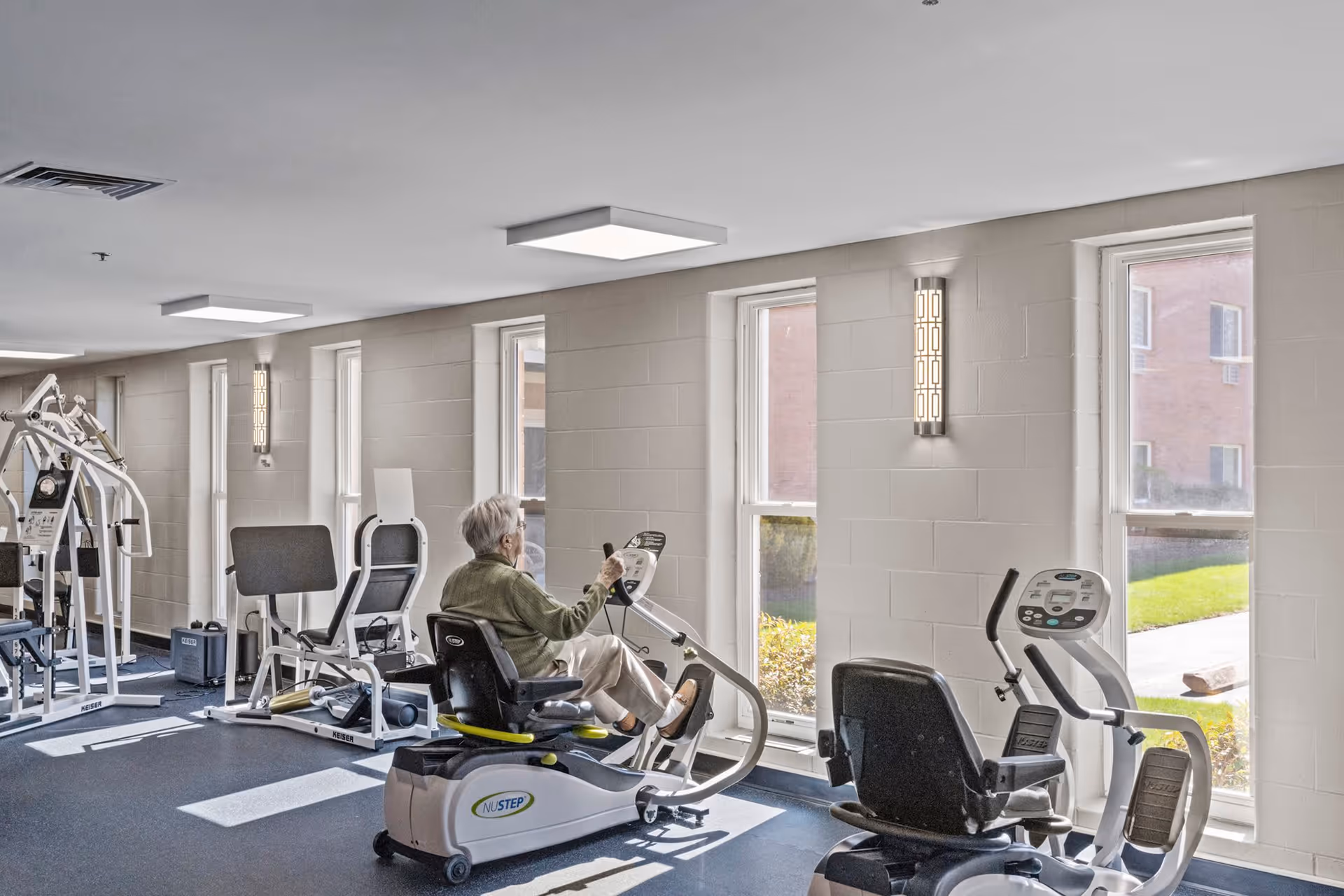 An elderly person exercising on a recumbent stationary bike in a bright fitness room with large windows letting in natural light. The room contains various exercise machines and equipment along the wall.