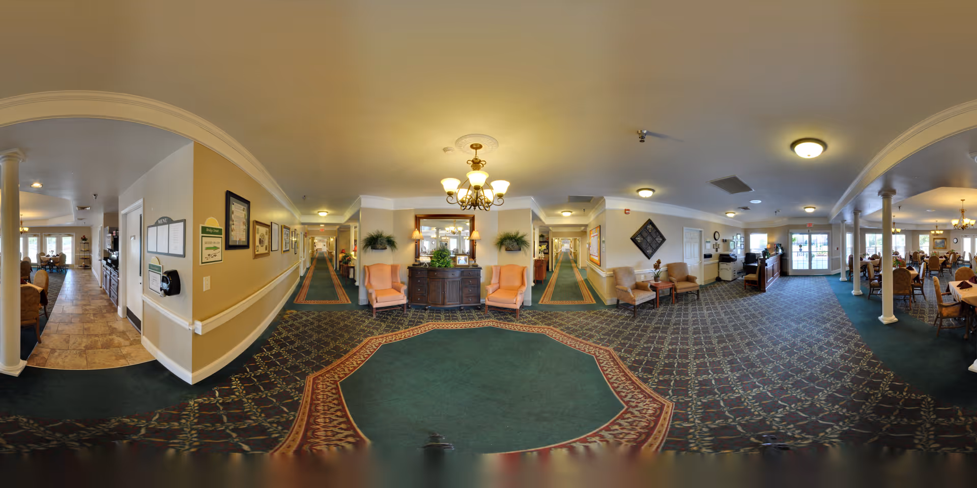 Panoramic interior view of a senior living facility lobby and common area with seating, a reception desk, and a dining room.
