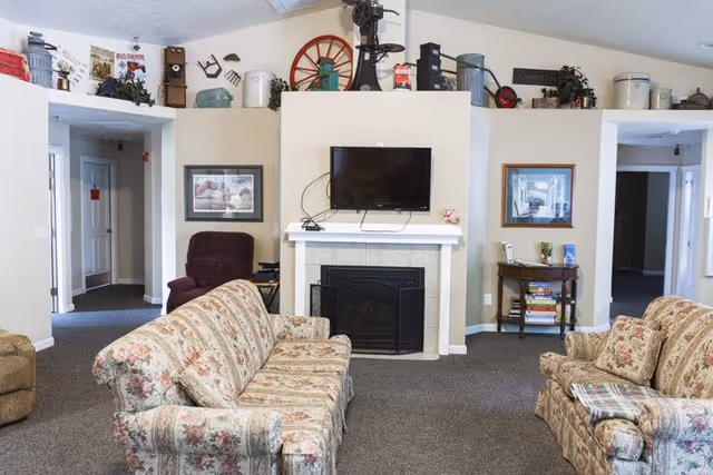 Cozy common living room with floral sofas facing a fireplace and wall-mounted TV, decorated with vintage items along a high shelf.