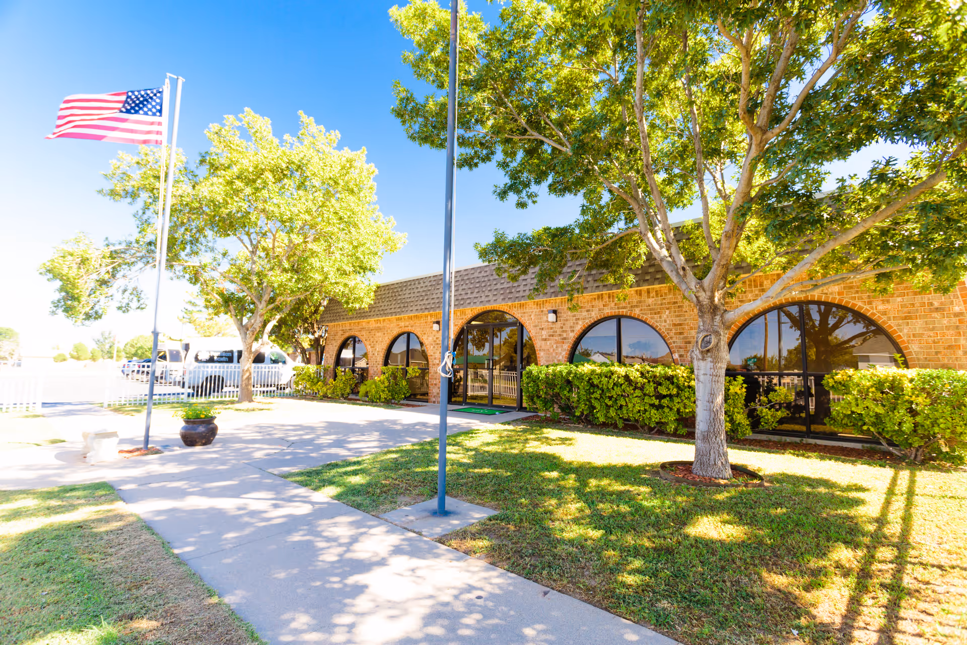 Exterior view of a single-story brick building with large arched windows, surrounded by green bushes and trees. An American flag is flying on a flagpole near the sidewalk leading to the entrance. The sky is clear and blue.