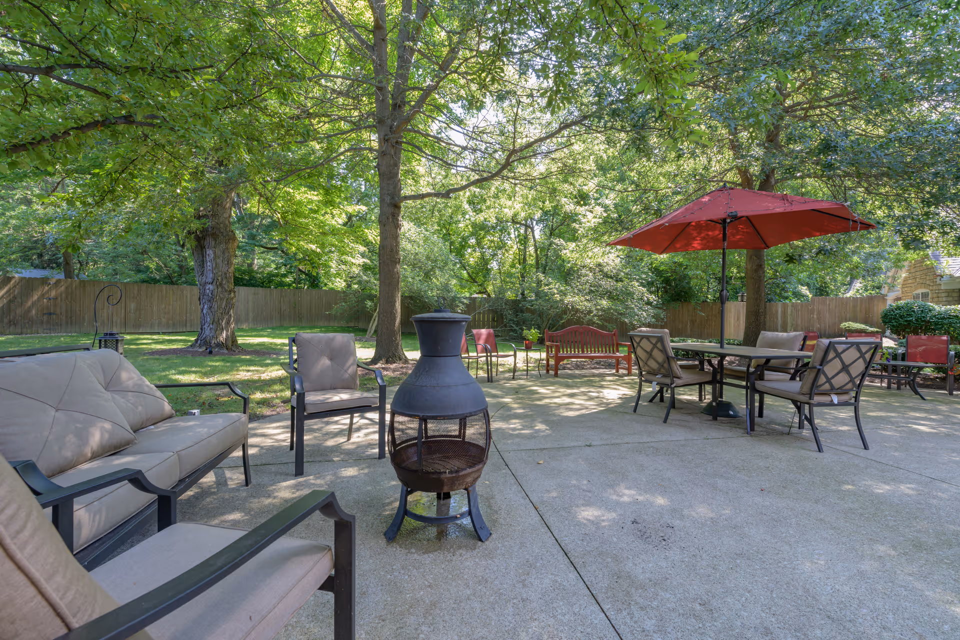 Outdoor patio area with cushioned chairs, a small fire pit, a table with a red umbrella, and benches surrounded by trees and a wooden fence.