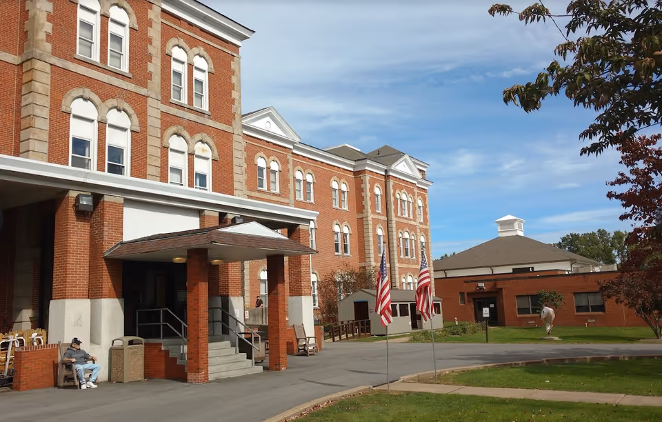 Exterior view of a large brick building with multiple windows and a covered entrance. Two American flags are displayed on flagpoles near the entrance. There is a person sitting on a bench near the building, and another building is visible in the background with a small sculpture on the lawn. Trees with autumn-colored leaves frame the scene under a partly cloudy sky.