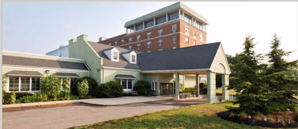 Exterior view of a senior living facility named Oasis at the Crescent, showing a light green building with a covered entrance and a taller brick building in the background, surrounded by greenery and trees.