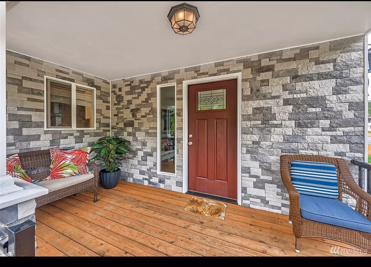 Covered porch area with wooden floor, stone-patterned walls, a red front door with decorative glass panel, a wicker loveseat with colorful tropical cushions, a potted plant, and a wicker chair with blue cushions.
