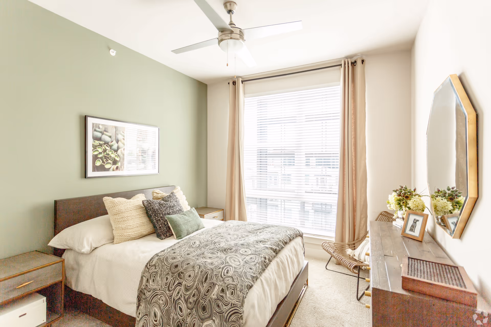 Sunlit bedroom with a bed against a green accent wall, bedside tables, a dresser with mirror and decor, and a large window with curtains.