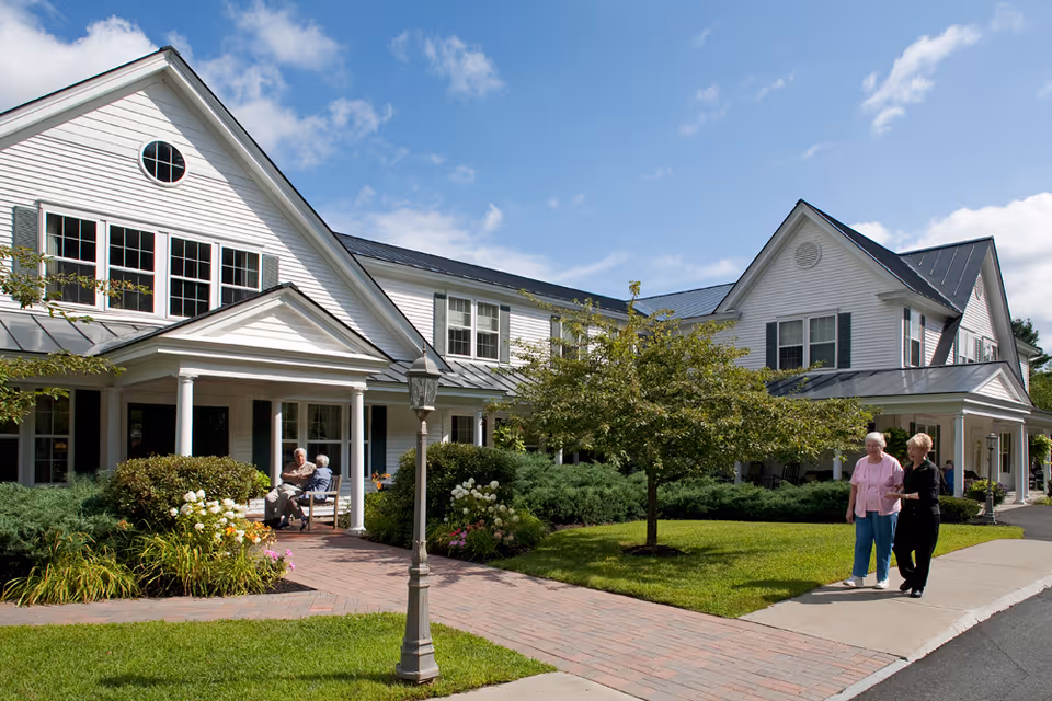 Exterior view of a white two-story senior living facility with green shutters and a metal roof. Two elderly people are sitting and talking on a porch with white columns, while two women walk on a sidewalk nearby. The area is landscaped with green bushes, a small tree, and flowers under a partly cloudy blue sky.