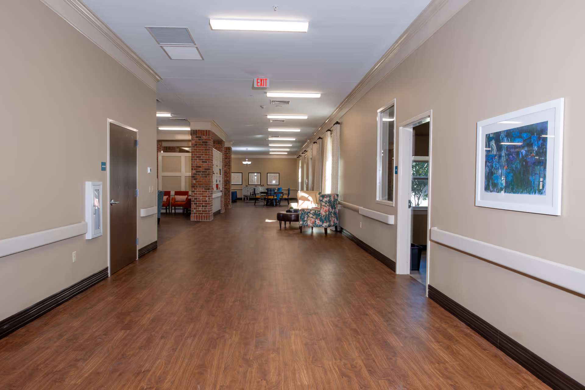 A wide hallway in an assisted living facility with wood flooring, beige walls, and ceiling lights. The hallway features handrails on both sides, a floral armchair, a small ottoman, and framed artwork on the walls. At the far end, there are tables and chairs arranged in a common area with brick pillars and windows letting in natural light.