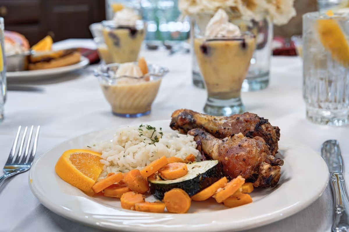 A plate of food on a white tablecloth featuring two grilled chicken drumsticks, white rice garnished with herbs, cooked carrots and zucchini, and a slice of orange. In the background, there are glasses of water with lemon and dessert cups with whipped cream.