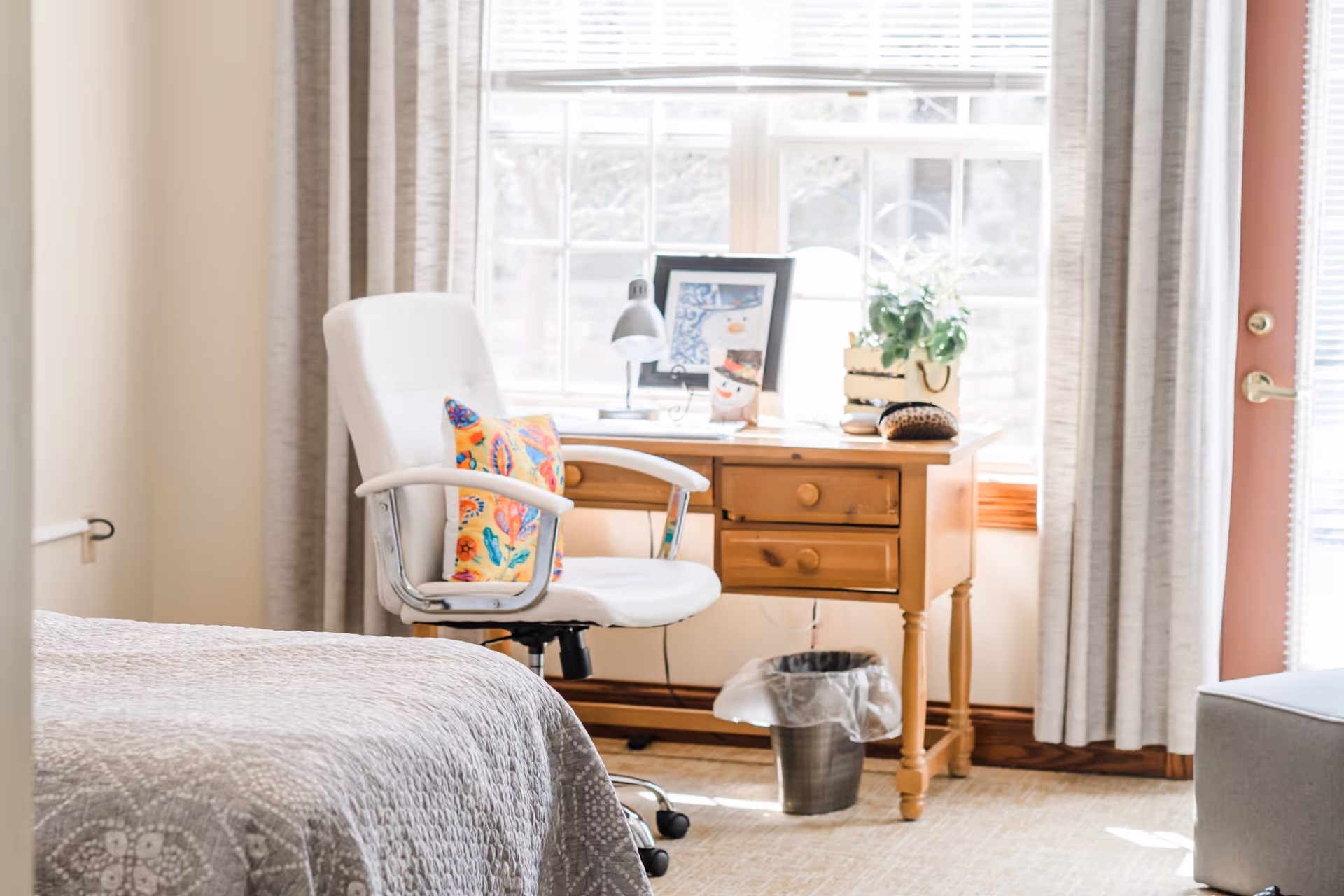 A bright room with a wooden desk placed in front of a window with white blinds and beige curtains. On the desk are a small plant, a desk lamp, and framed pictures. In front of the desk is a white office chair with a colorful cushion. Part of a bed with a gray patterned bedspread is visible on the left side, and a door with a window and beige curtains is on the right side. A small trash can is under the desk.