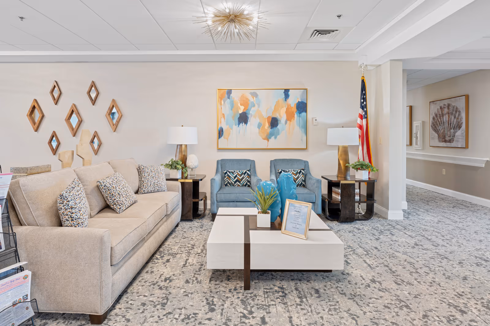 A bright and modern living room area featuring a beige sofa with patterned cushions, two blue armchairs with decorative pillows, a white coffee table with plants and framed artwork, two side tables with lamps, a wall decorated with diamond-shaped mirrors, and an American flag in the corner. The room has a patterned carpet and light-colored walls with abstract and shell-themed artwork.