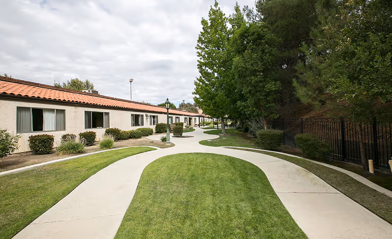 Curved concrete pathways surrounded by green grass and trees leading alongside a single-story building with a tiled roof and multiple windows under a cloudy sky.
