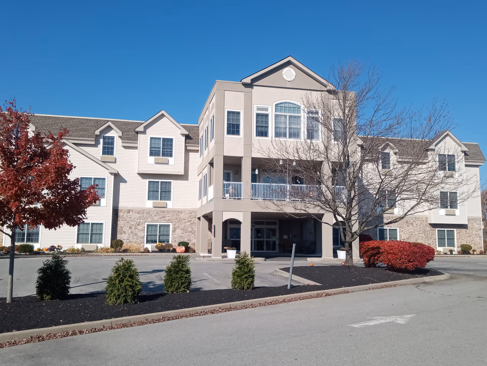 Three-story beige and stone senior living building with a covered entrance, balcony, and landscaped parking area under a clear blue sky.