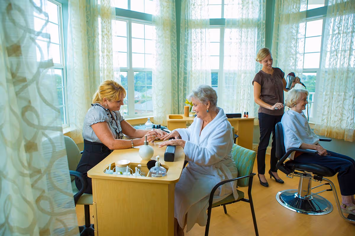 A bright room with large windows and sheer curtains where an elderly woman in a white robe is receiving a manicure from a woman seated at a wooden table. Another elderly woman, also in a white robe, is seated in a salon chair while a stylist blow-dries her hair. The room has a warm and welcoming atmosphere.