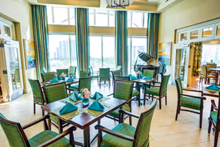 Bright dining room with multiple tables set with green napkins and floral centerpieces, green cushioned chairs, large windows with curtains letting in natural light, and a grand piano in the corner.