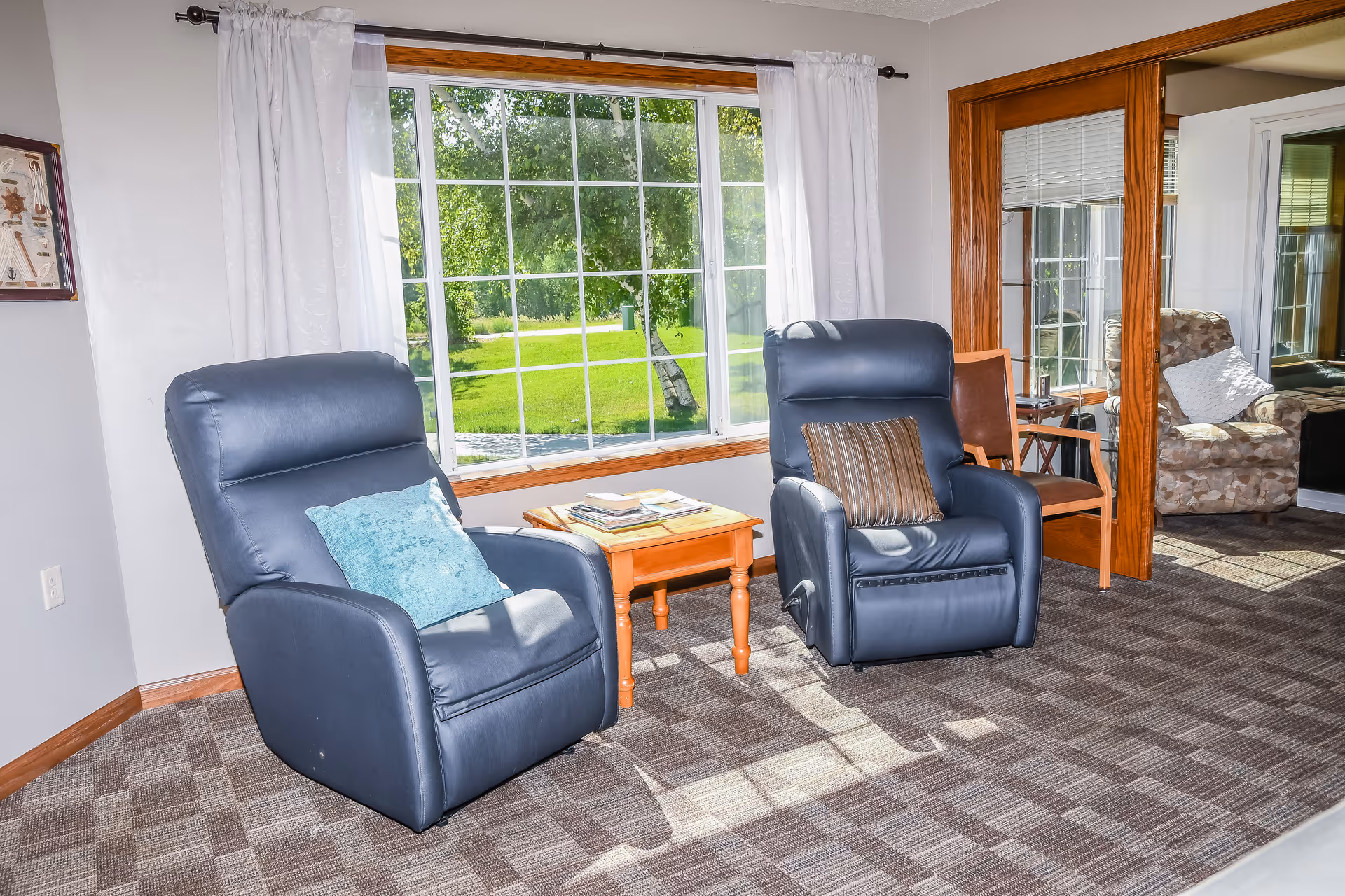 Two blue recliner chairs and a small wooden side table in a bright seating area with a large window showing a green lawn outside.