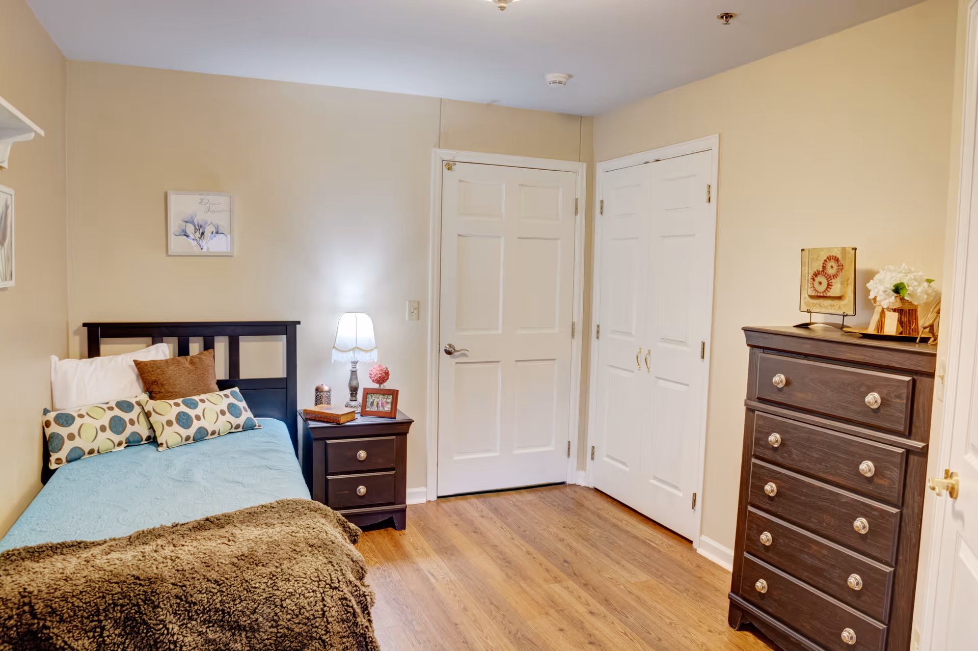 A cozy bedroom with a single bed featuring a blue bedspread, multiple pillows, and a brown throw blanket. Next to the bed is a dark wooden nightstand with a lamp, a small decorative item, and a framed photo. The room has light beige walls, wooden flooring, a dark wooden dresser with decorative items on top, and two white doors, one of which appears to be a closet.