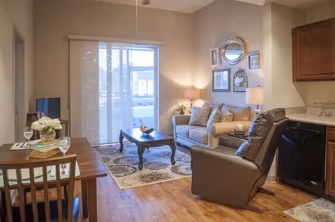 A cozy living room area with a beige sofa, a dark brown recliner chair, a wooden coffee table on a patterned rug, and a small dining table with two chairs. The room has wooden flooring, a sliding glass door with vertical blinds leading outside, and a kitchen area with wooden cabinets and a dishwasher. The walls are decorated with framed pictures and a round mirror.