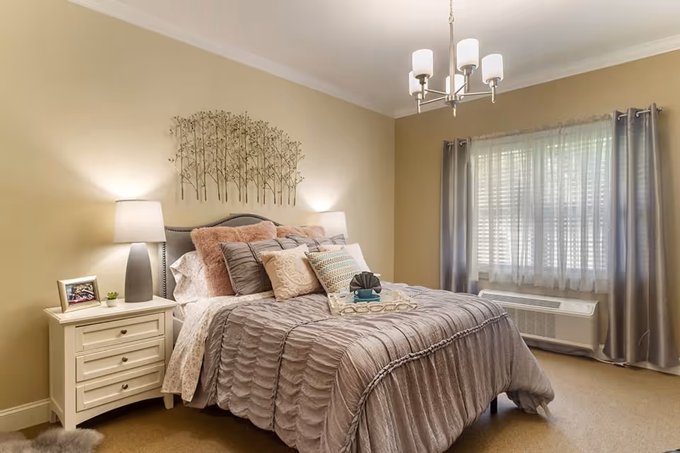 A cozy bedroom with a neatly made bed featuring multiple pillows and a textured gray comforter. On the left side of the bed is a white nightstand with a lamp, a small plant, and a framed photo. Above the bed is a decorative metal wall art piece depicting trees. A window with sheer white curtains and gray drapes is on the right wall, with an air conditioning unit below it. A modern chandelier hangs from the ceiling.
