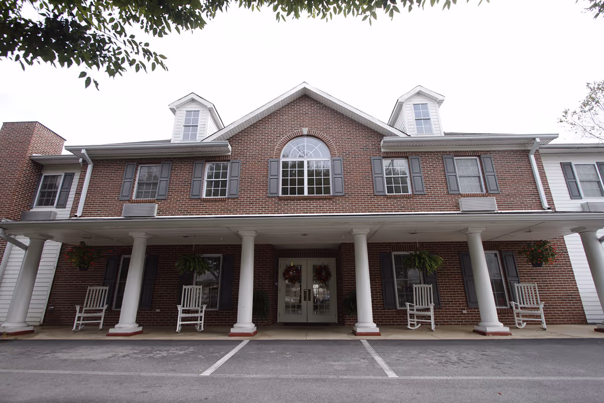 Front exterior view of a two-story brick building with white columns supporting a covered porch. The building has multiple windows with gray shutters, two dormer windows on the roof, and a central arched window above the entrance. Four white rocking chairs and hanging plants are placed on the porch. The parking area in front has marked spaces.