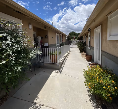 Outdoor walkway between beige assisted-living units with potted plants and chairs under a cloudy sky.