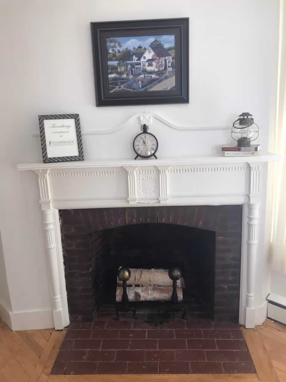 White decorative mantel and brick fireplace with a framed painting above, a clock and small decorative items on the mantel.