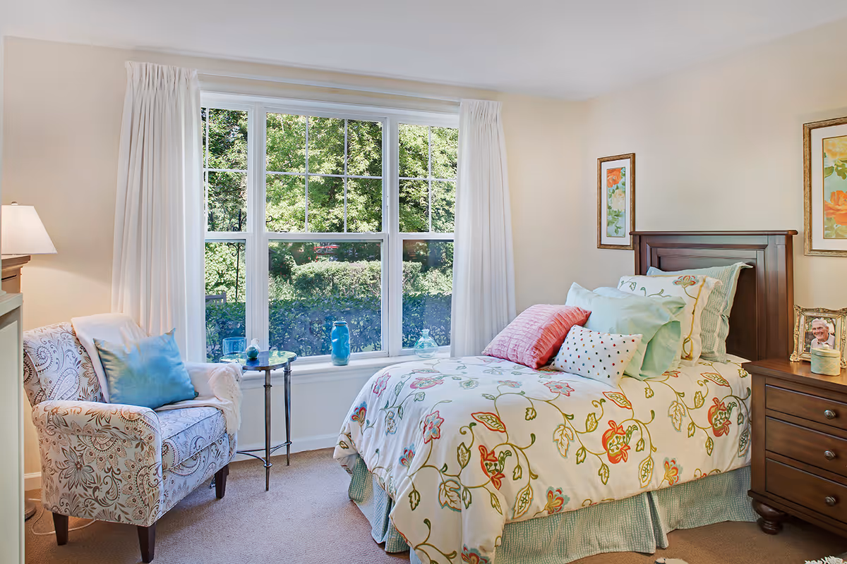 A cozy bedroom with a single bed featuring floral bedding and multiple pillows. Next to the bed is a wooden nightstand with a framed photo and a small container. A patterned armchair with a blue pillow and a white throw blanket sits beside a small round glass table. Large windows with white curtains let in natural light and show green foliage outside.