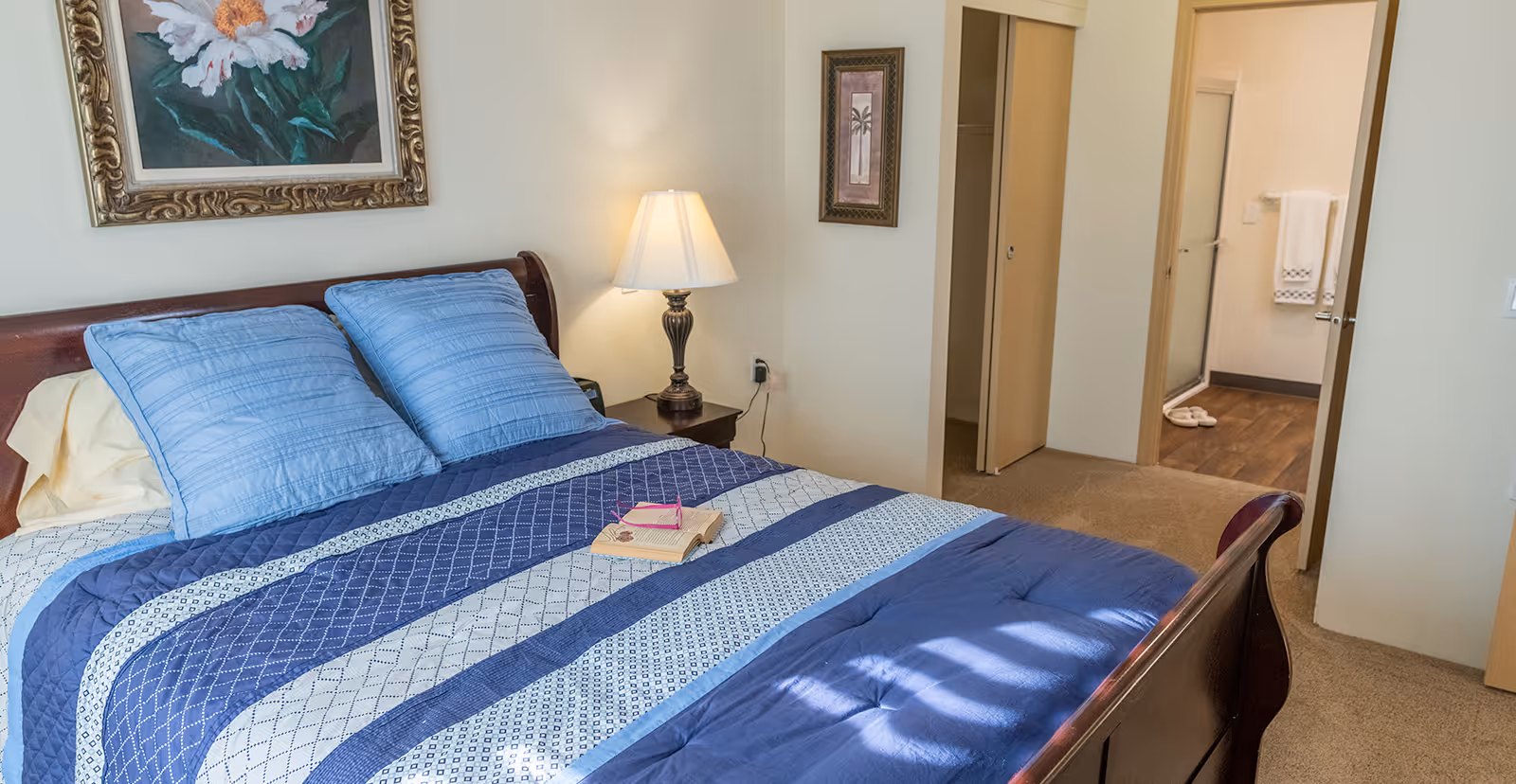 A bedroom with a wooden bed frame, blue and white striped bedding, and two blue pillows. A nightstand with a lamp is beside the bed. There are two framed pictures on the walls. An open door reveals a bathroom with a shower, towel rack, and slippers on the floor.