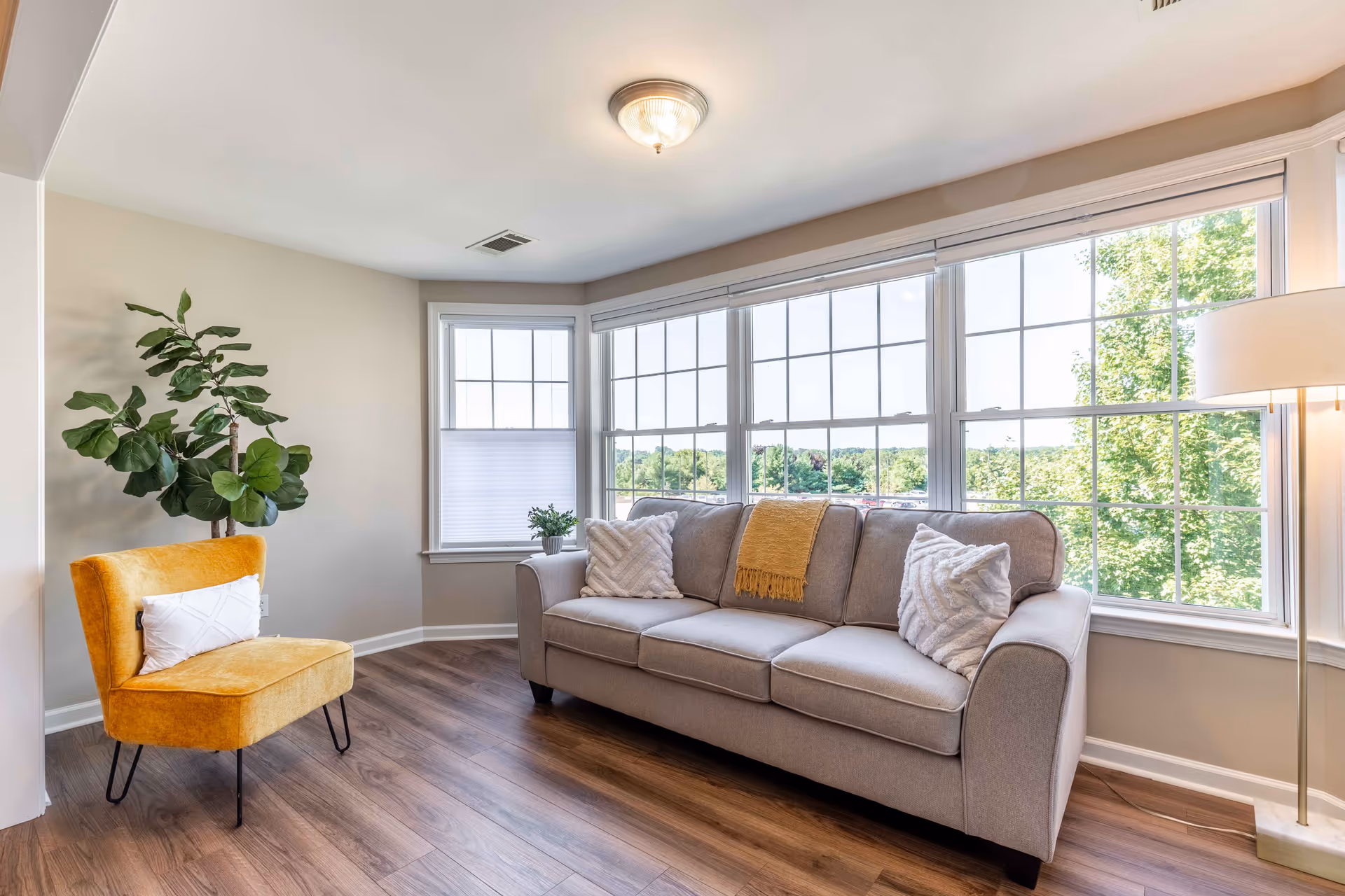A bright living room with large windows showing a green outdoor view. The room features a gray sofa with two white pillows and a yellow throw blanket, a yellow upholstered chair with a white pillow, a tall floor lamp, and a potted plant in the corner. The floor is wood with a light finish.