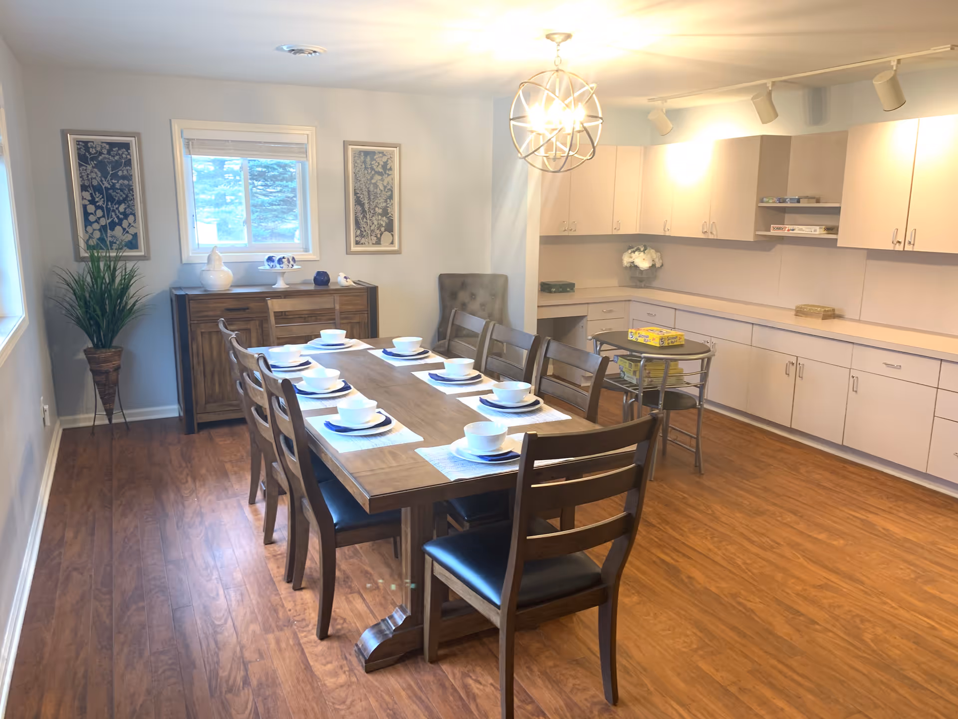 A dining room with a long wooden table set for eight people with white bowls and plates on placemats. The room has wooden flooring, a sideboard with decorative items, two framed artworks on the wall, a potted plant, and a modern chandelier hanging from the ceiling. There are white cabinets along one wall with a small round table and chairs in front of them.