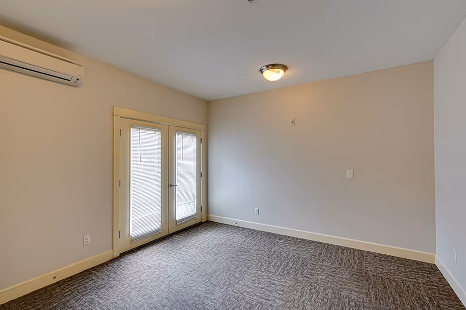 Empty room with beige walls, carpeted floor, a ceiling light fixture, a wall-mounted air conditioning unit, and double glass doors with blinds.
