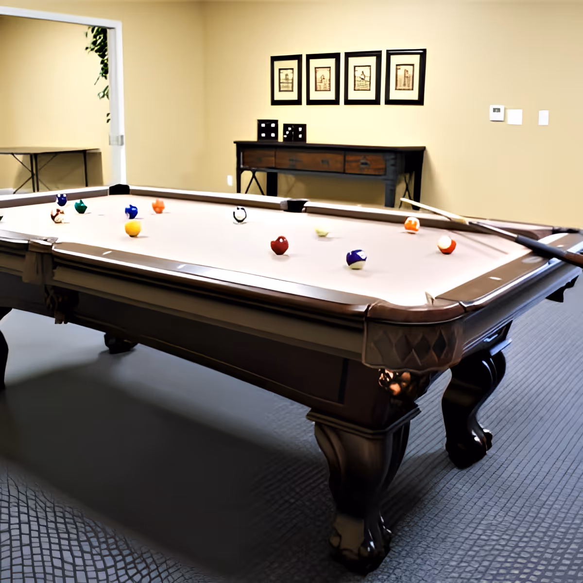 A pool table with billiard balls arranged on it in a room with beige walls. There is a wooden console table against the wall with framed artwork above it and a doorway leading to another room on the left.
