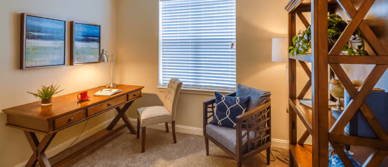 Cozy sitting and desk area with a wooden desk, upholstered chairs, bookshelf, window with blinds, and wall art.