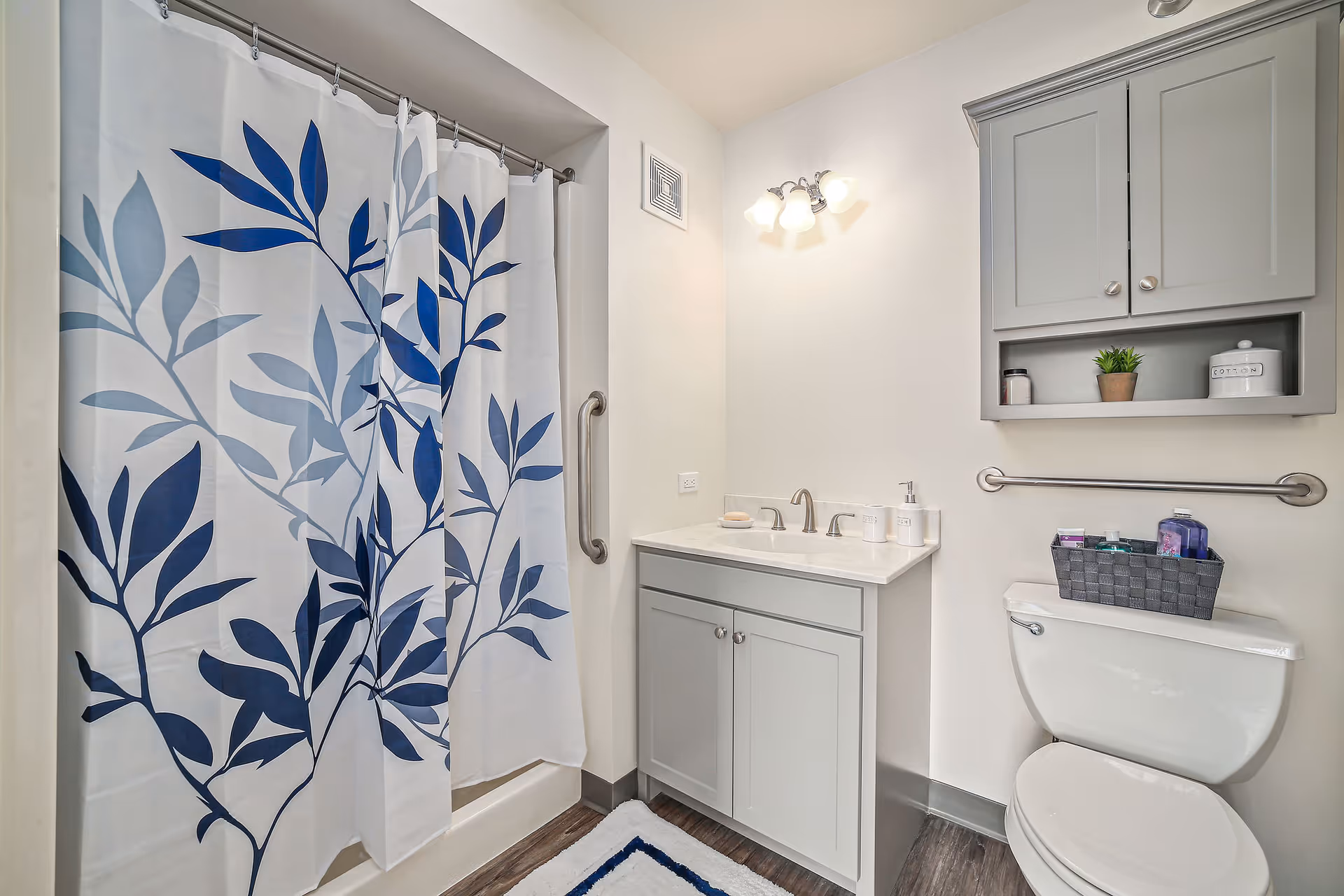 A clean and modern bathroom featuring a white shower curtain with blue leaf patterns, a white vanity with a sink and faucet, a wall-mounted cabinet with decorative items, a toilet with a basket of toiletries on top, and a grab bar next to the shower.
