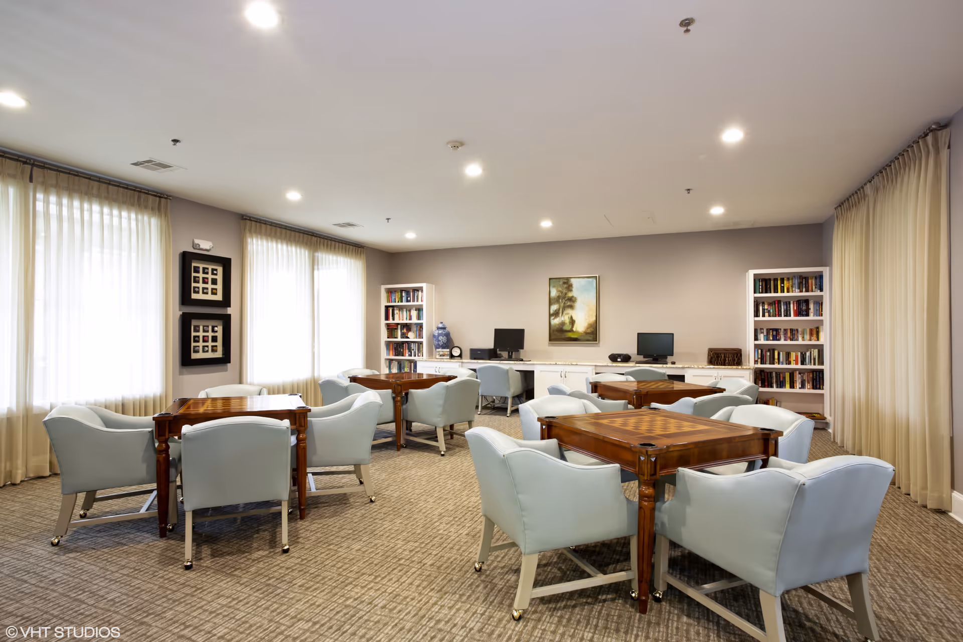 A well-lit common room with several wooden tables and light blue upholstered chairs arranged around them. The room has large windows with sheer curtains, bookshelves filled with books, two computer stations, and a framed painting on the wall.