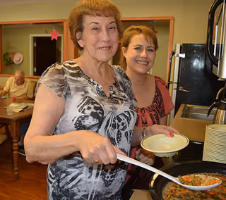 Two women smiling while serving food from a large pot in a dining area of a nursing and rehabilitation center. One woman is holding a ladle and the other is holding a plate. In the background, an elderly person is seated at a table.
