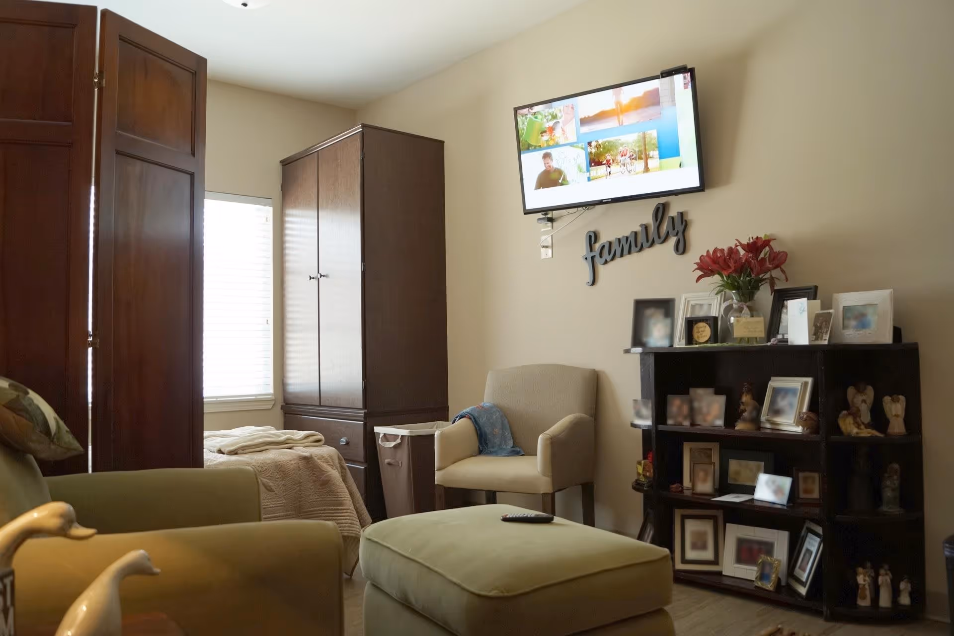 A cozy living room area with a green armchair and matching ottoman, a beige armchair with a blue blanket draped over it, a dark wooden cabinet filled with framed photos and decorative items, a wall-mounted TV displaying various images, and the word 'family' mounted on the wall beneath the TV. There is a window with blinds partially open and a bed with a beige blanket partially visible behind a wooden folding screen.