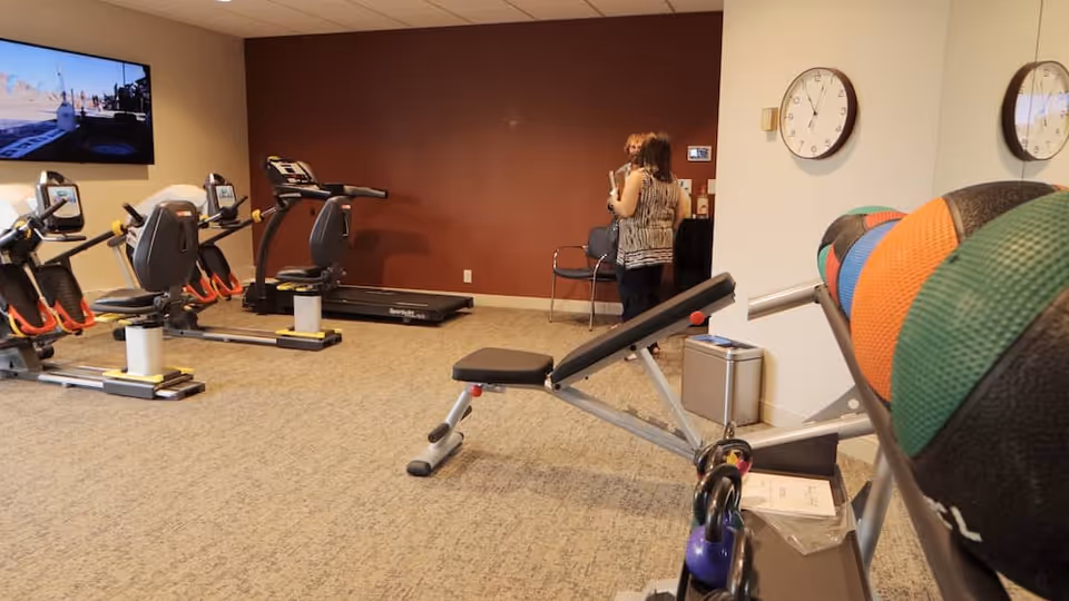 Indoor fitness room with exercise equipment including recumbent bikes, a treadmill, a weight bench, kettlebells, and colorful medicine balls. Two women are standing and talking near a chair against a brown wall. A large wall clock and a mounted TV are also visible.