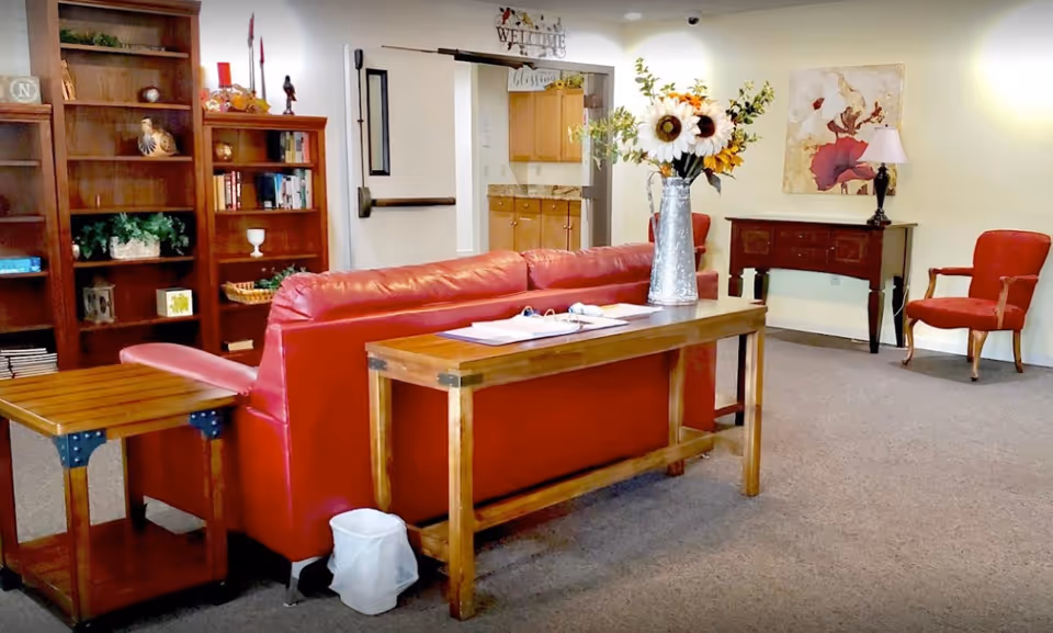 A cozy living room area with a red leather sofa, wooden side tables, and a wooden console table behind the sofa holding a vase with sunflowers. There are bookshelves with books and decorative items on the left, a red armchair on the right, a wooden sideboard with a lamp and artwork on the wall, and a doorway leading to a kitchen area in the background.