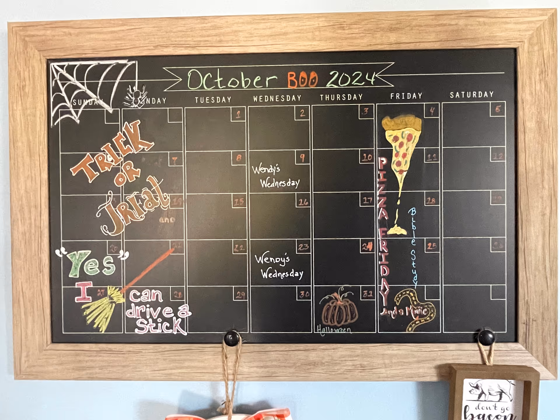 A wooden-framed black chalkboard calendar for October 2024 with colorful chalk drawings and writings. It features a spider web and spider in the top left corner, the words 'Trick or Treat' in orange and white, and a broomstick with the phrase 'Yes I can drive a stick' in green, red, and white. The calendar includes notes such as 'Wendy's Wednesday' on the 9th and 23rd, a slice of pizza drawing on the 4th, 'PIZZA FRIDAY' written vertically on the 4th, 'Bible Study' on the 25th, a pumpkin drawing on the 31st with 'Halloween' written underneath, and a snake drawing near the bottom right. The calendar is hanging on a wall with two hooks, one of which holds a small wooden sign with a pig illustration and the phrase 'don't go bacon my heart'.