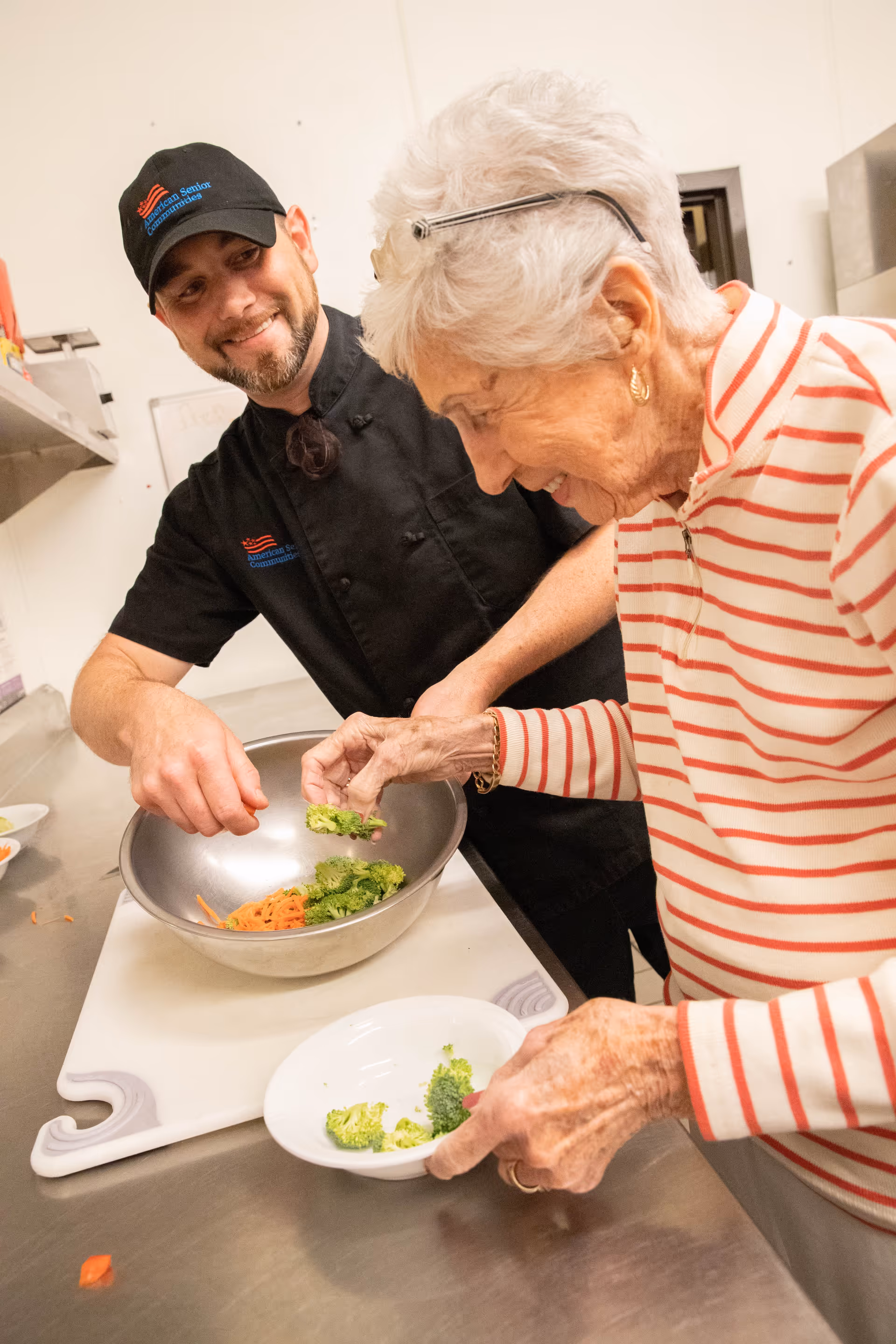 A chef and an older woman preparing broccoli and shredded carrots in a commercial kitchen bowl.