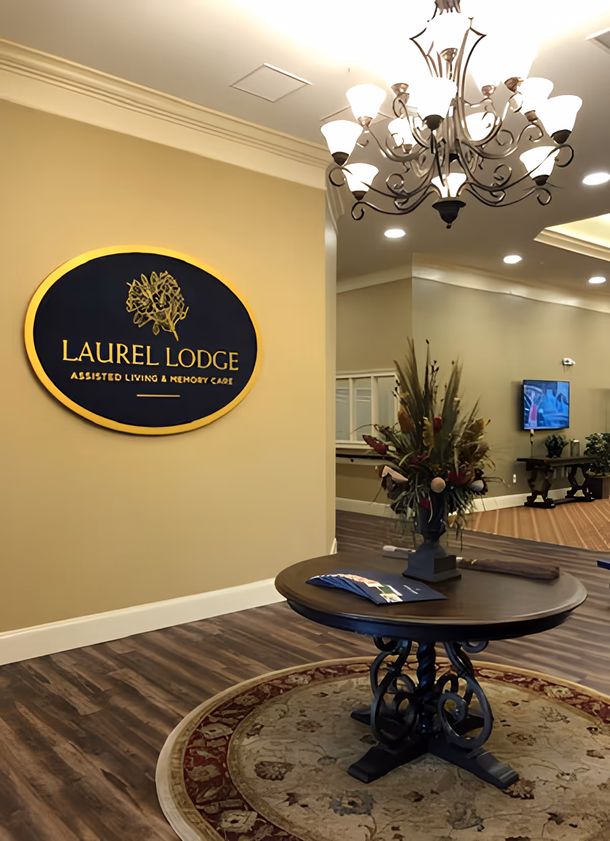 Lobby interior of Laurel Lodge Assisted Living & Memory Care showing a wall sign, round table with a floral arrangement on a rug, and a chandelier.