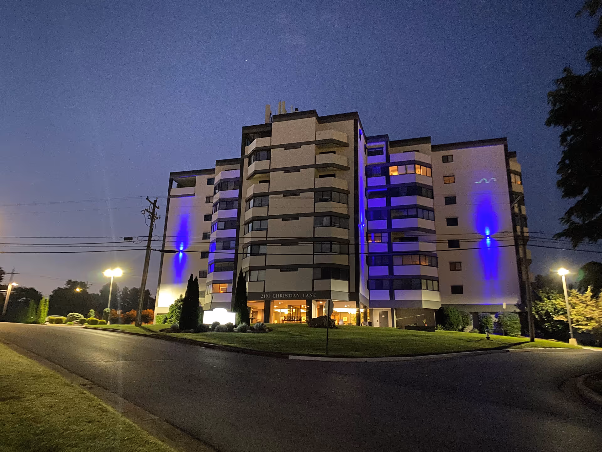 Exterior nighttime view of a multi-story senior living facility building with blue lights illuminating parts of the facade. The building is surrounded by a well-maintained lawn and trees, with streetlights and a paved road in the foreground.