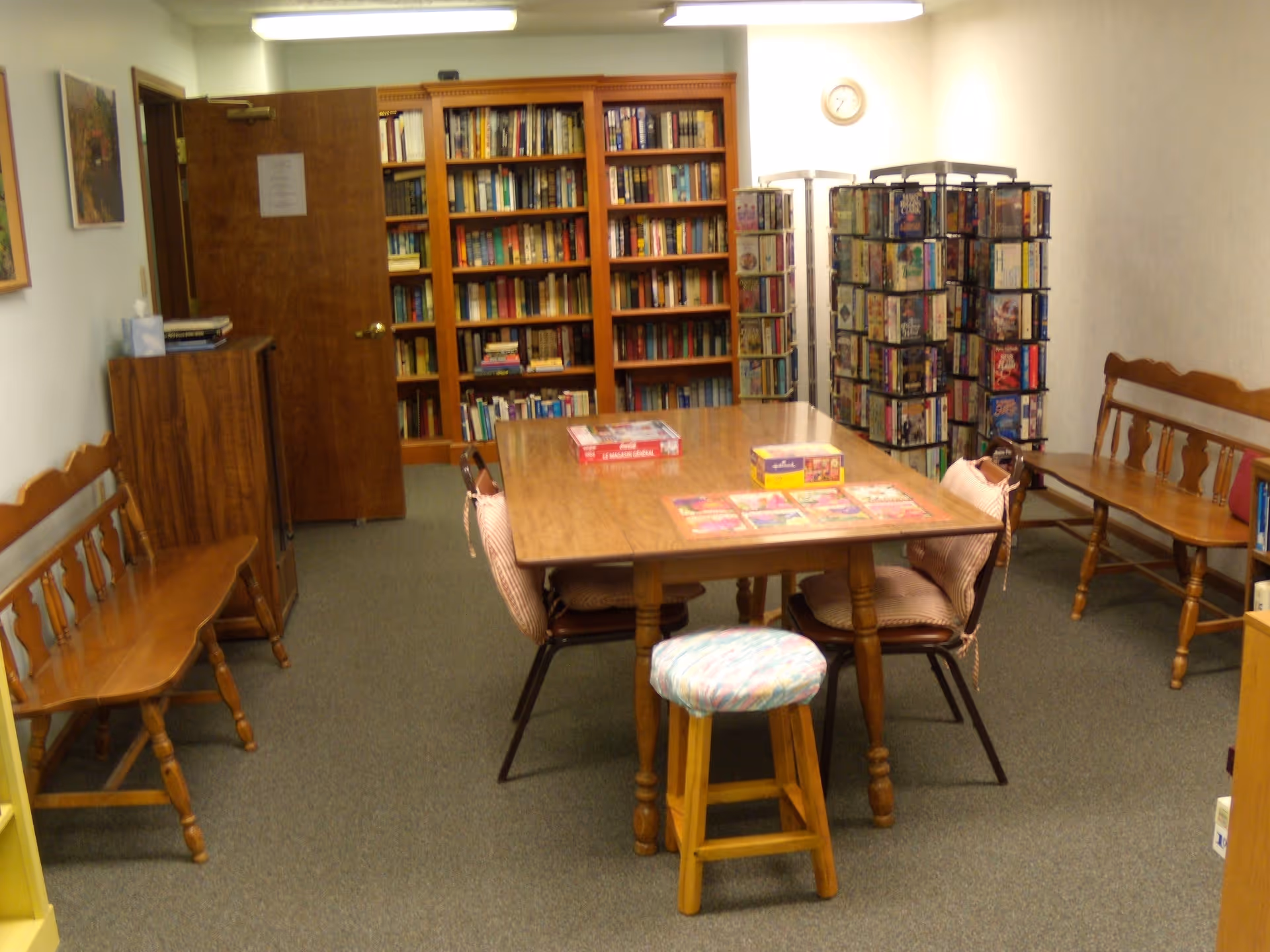A cozy room with a wooden table surrounded by chairs and a cushioned stool. The table has board games and puzzles on it. There are wooden benches along the walls, a bookshelf filled with books, and rotating racks with DVDs or similar media. The room has carpeted flooring and light-colored walls with a clock and framed pictures.