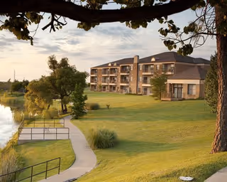 Three-story residential building with balconies overlooking a pond, a curved walkway and a broad grassy lawn under a cloudy sky.