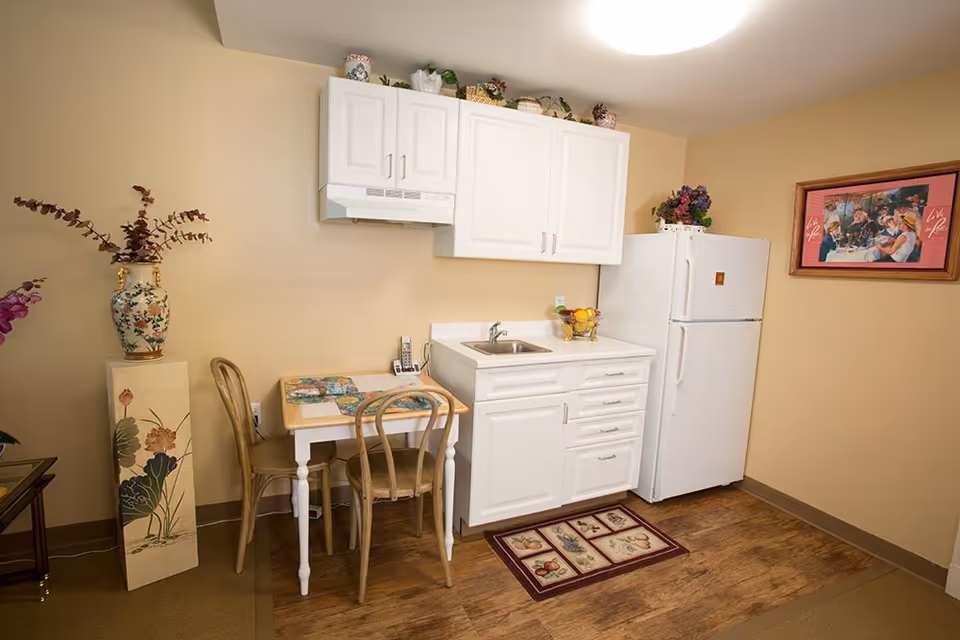 Small kitchen area with white cabinets, a white refrigerator, a small sink, and a two-chair dining table with placemats. There is a decorative vase with flowers on a stand to the left and a framed picture on the beige wall to the right.