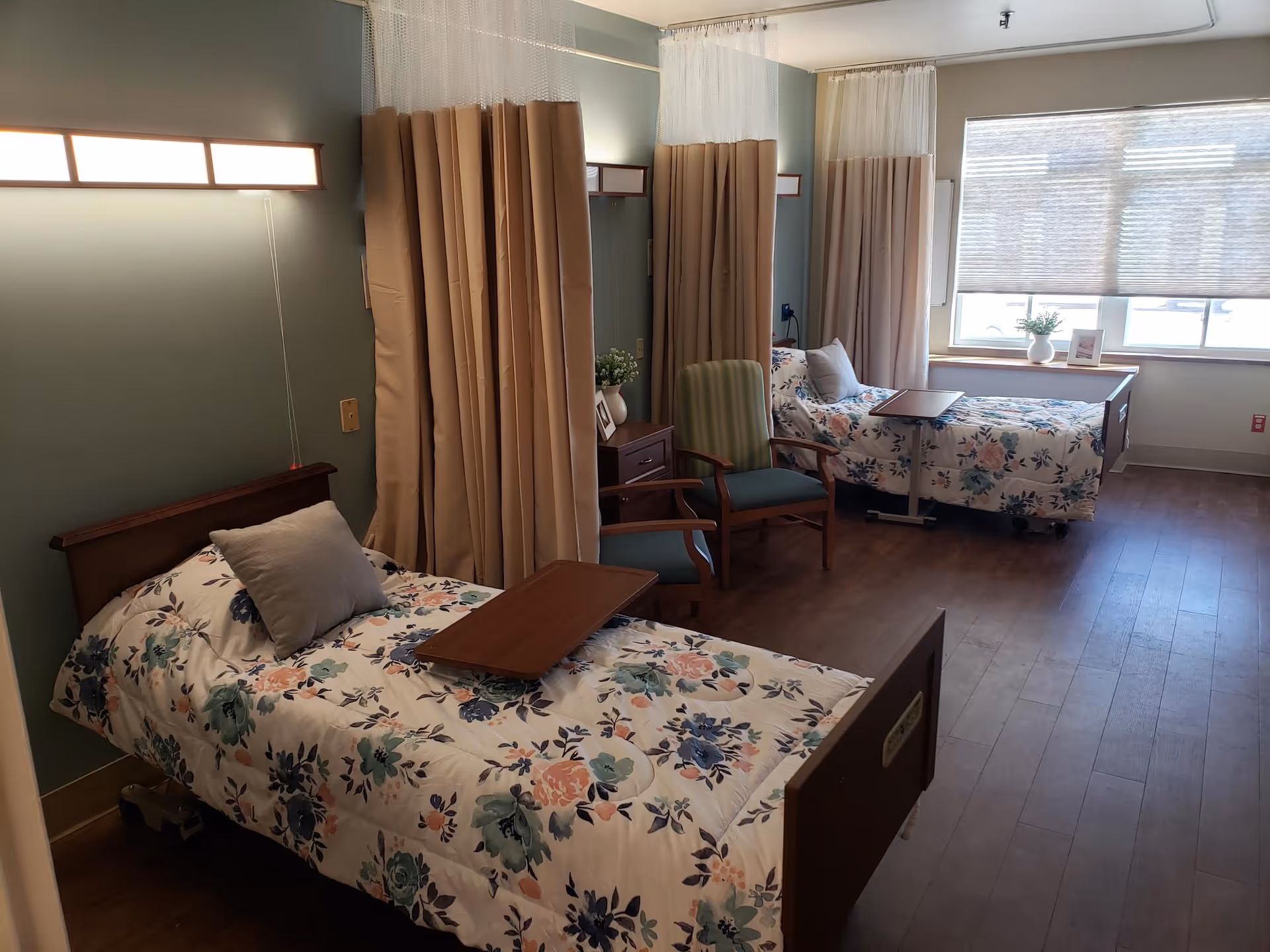 A shared bedroom in a healthcare facility with two beds separated by privacy curtains. Each bed has floral bedding and a pillow, with a wooden tray table on top. There are two chairs between the beds and a window with blinds letting in natural light. A small table with a plant and picture frame is placed near the window.